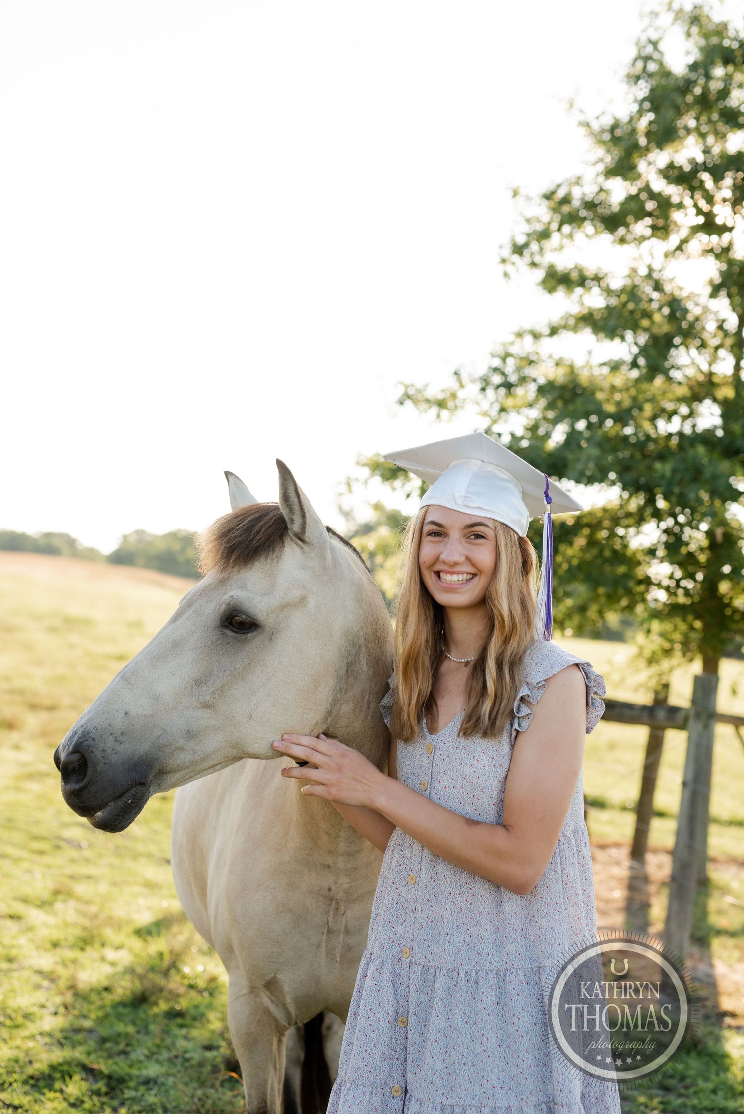 A graduate stands beside a white horse in a sunny field with wooden fencing.