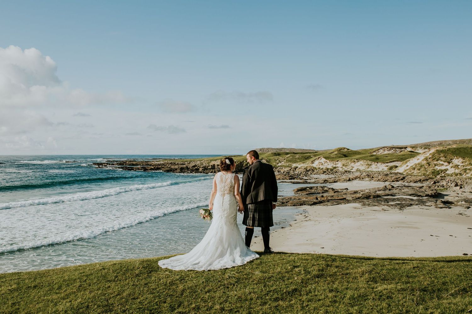 A wedding couple stands together on a grassy cliff overlooking a dramatic coastal beach landscape.