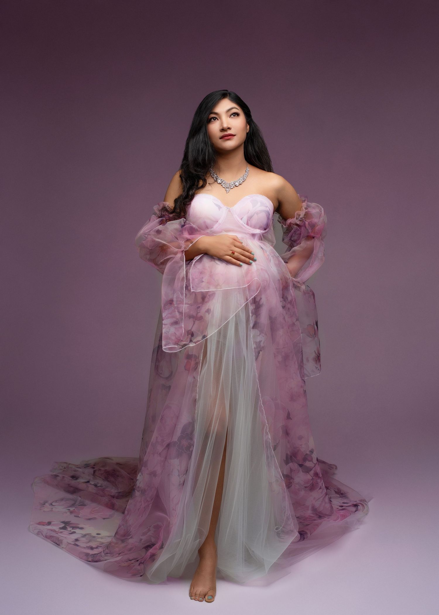an expecting mother lifts her eyes to the sky with one hand on her belly wearing a purple flowery dress