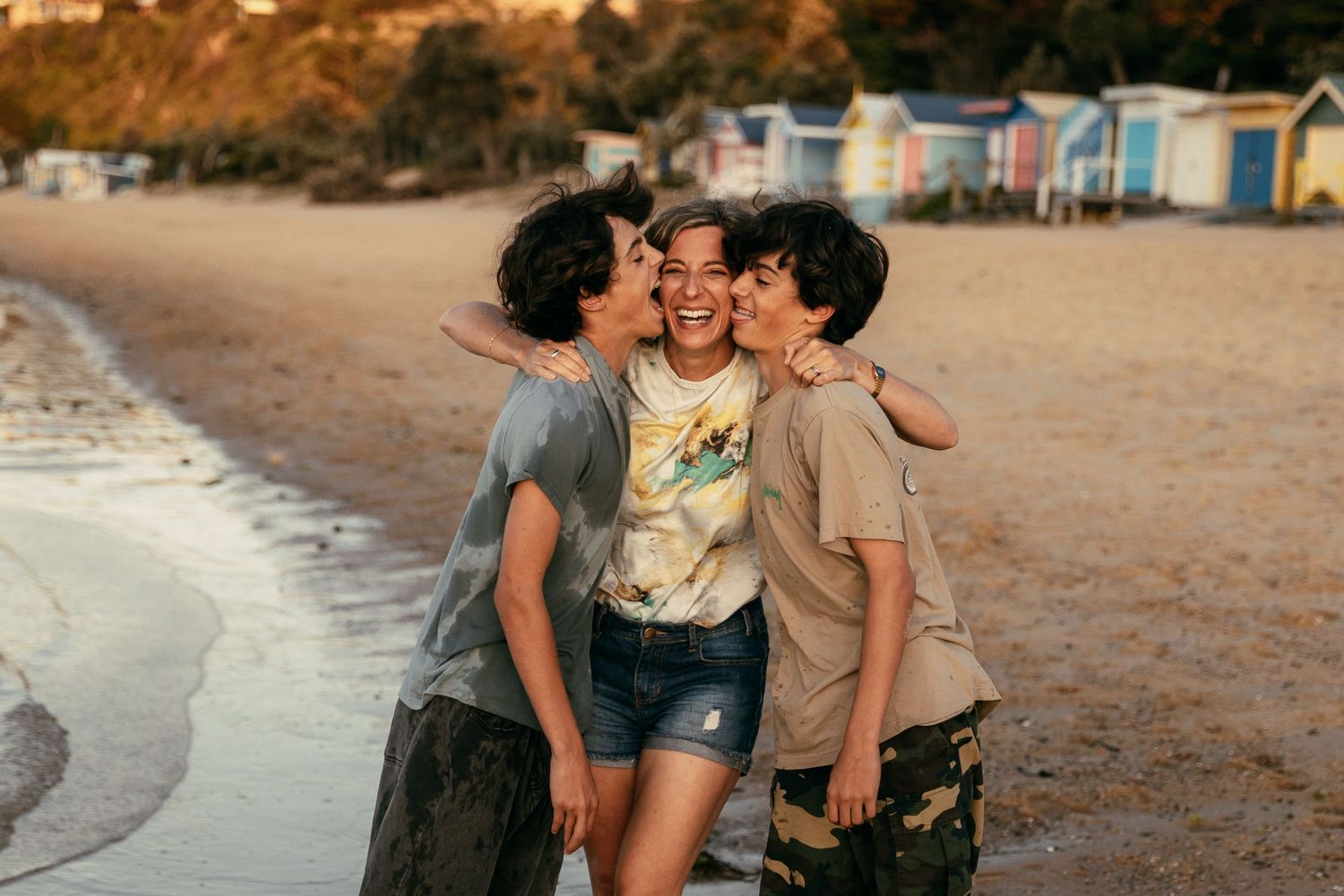 teenage boys kissing mum on cheek on beach with beach huts in background