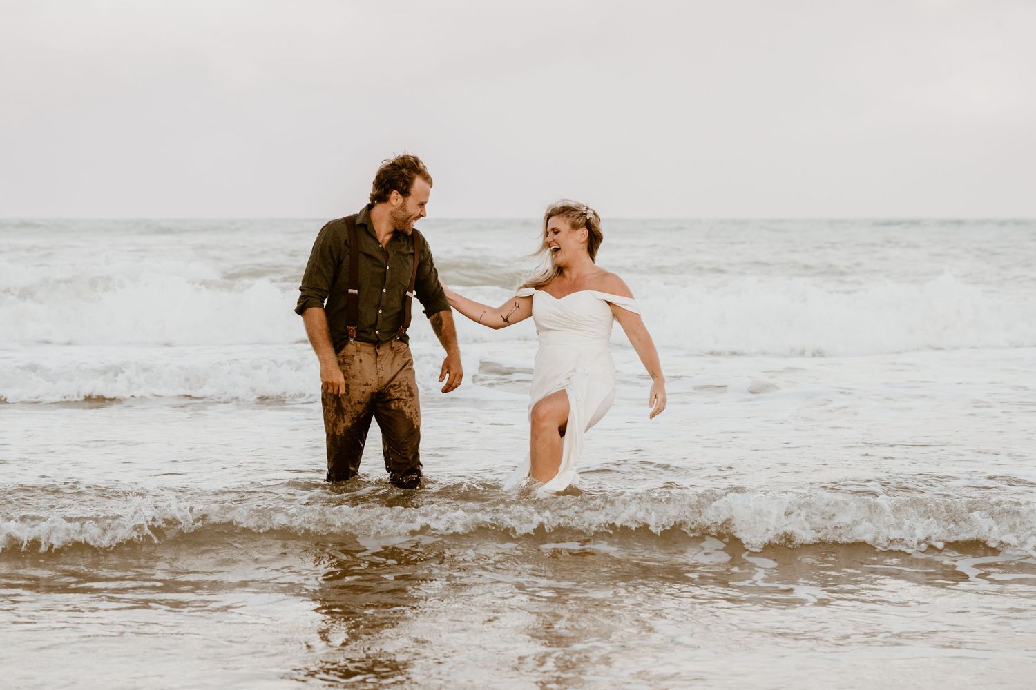 A couple playing in ocean waves wearing wedding attire.