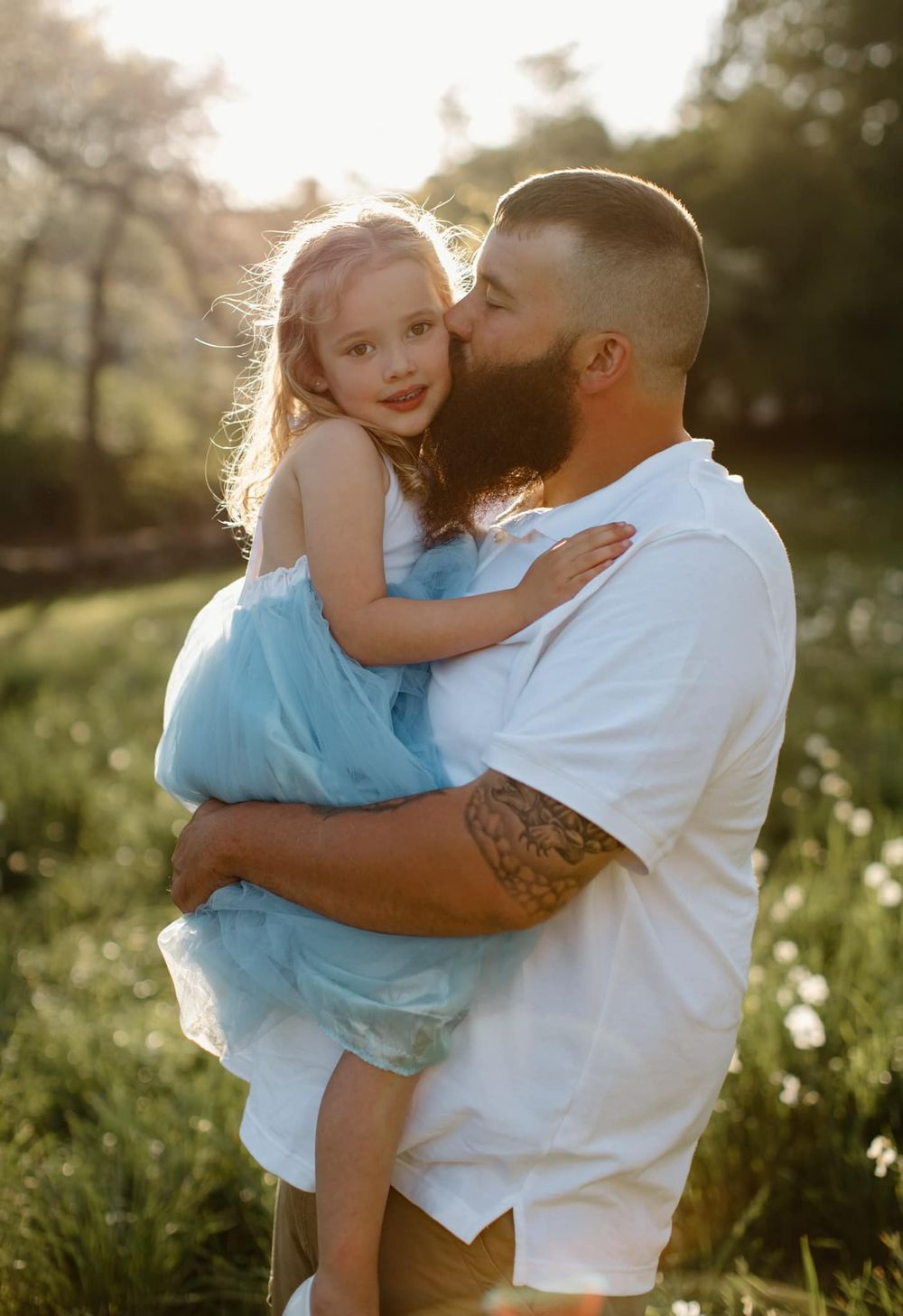 Father and child share tender moments in a sunlit field wearing white and blue outfits.