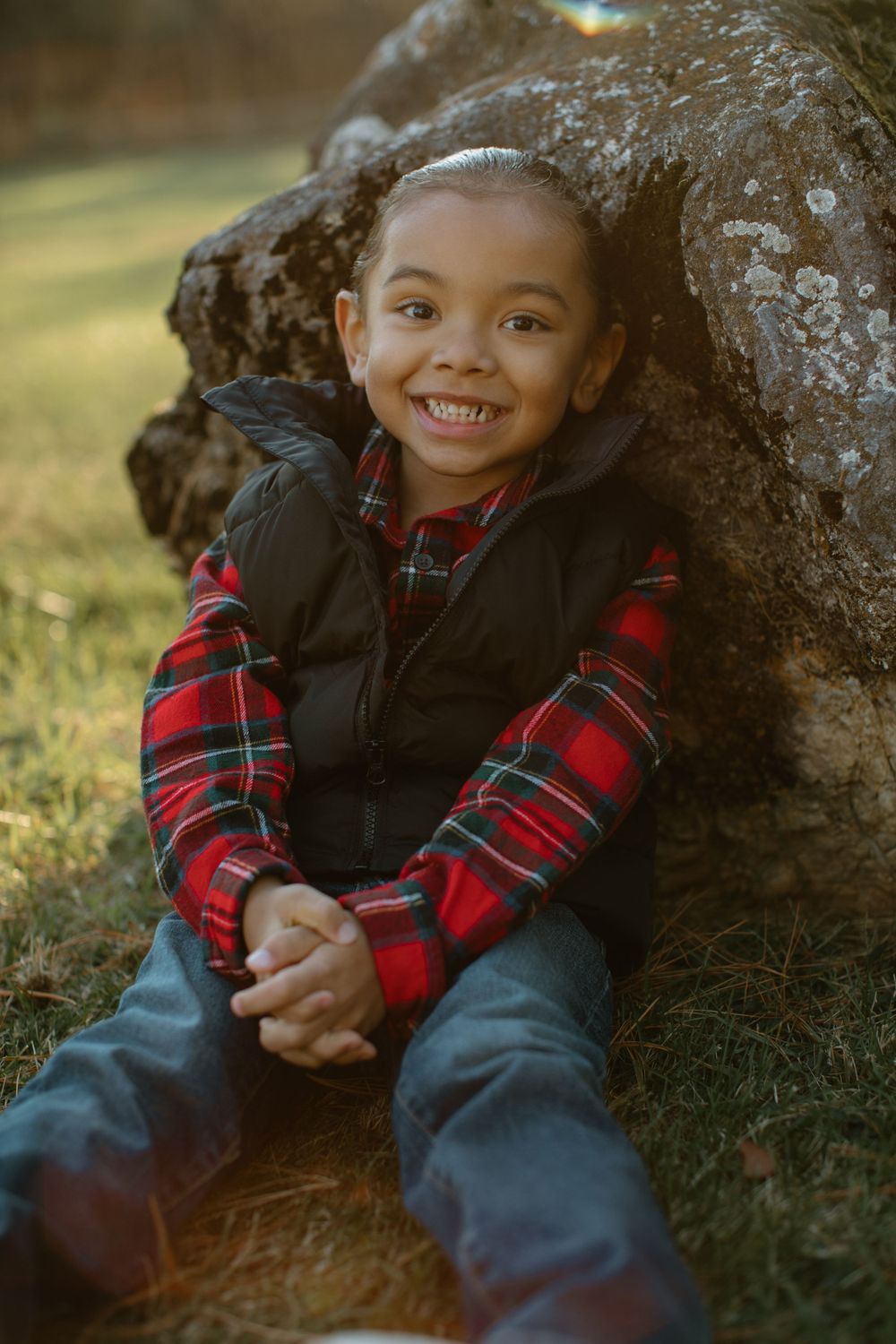 Child in plaid and black jacket sits against rock formation outdoors.