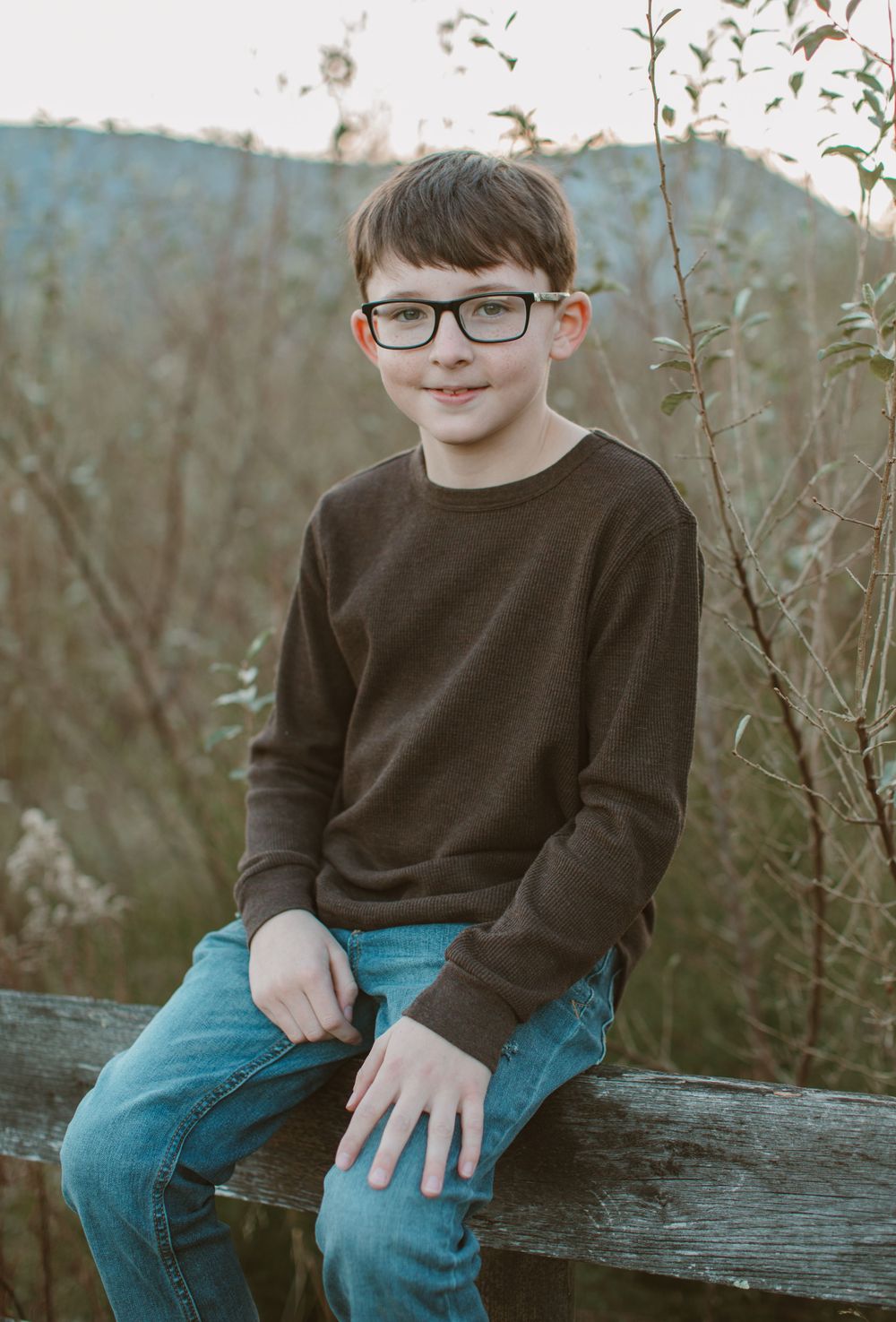 Child in brown sweater and jeans sits on wooden bench outdoors.