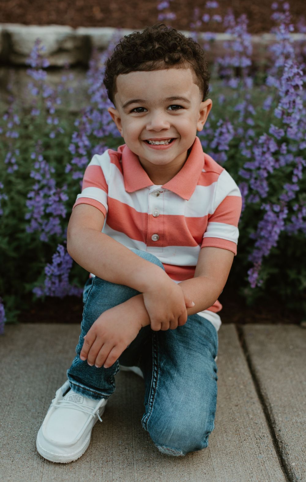 Child in striped polo shirt smiles while sitting by purple flowers.