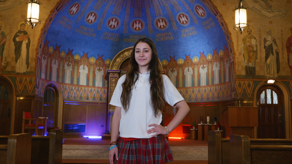 Student in white shirt and plaid skirt standing in ornate church interior with blue dome ceiling.