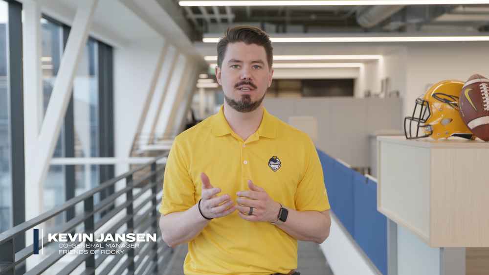 Person in white polo shirt speaking in modern office setting with football helmet in background.