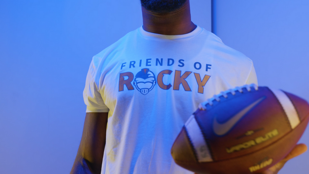 Person holding a football wearing a white 'Friends of Rocky' t-shirt in blue lighting.