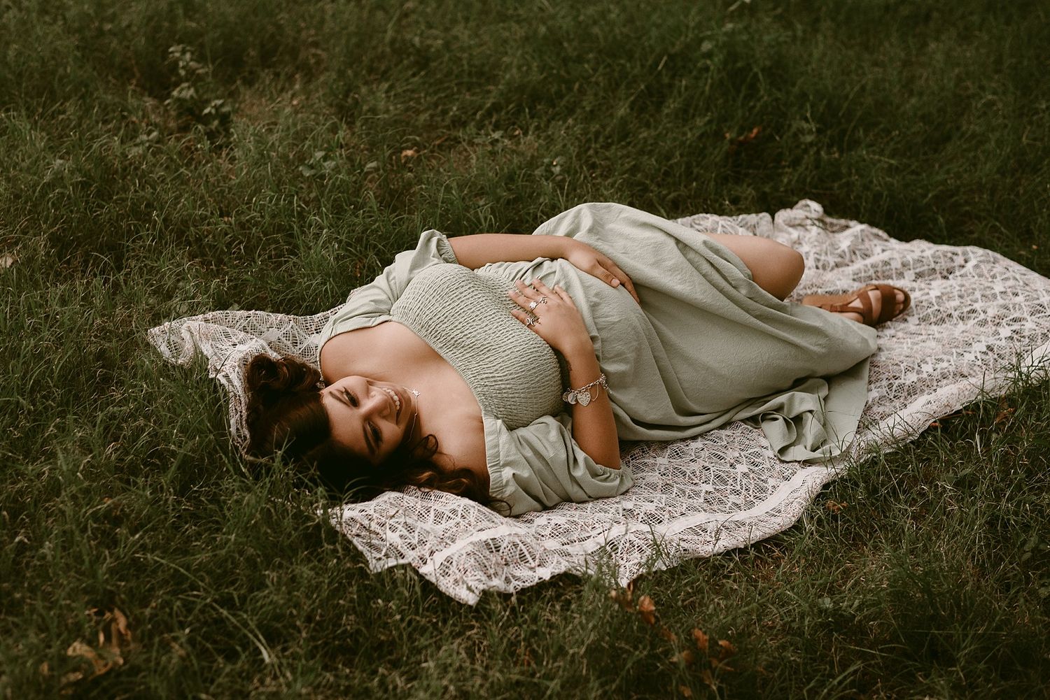 Artistic maternity photo of woman in grey dress on lace blanket outdoors.