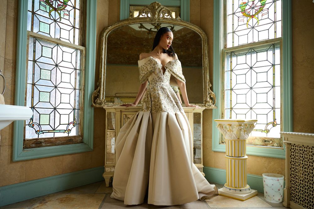 Model in ethereal vintage-style cream dress poses elegantly between ornate windows in elegant interior setting.