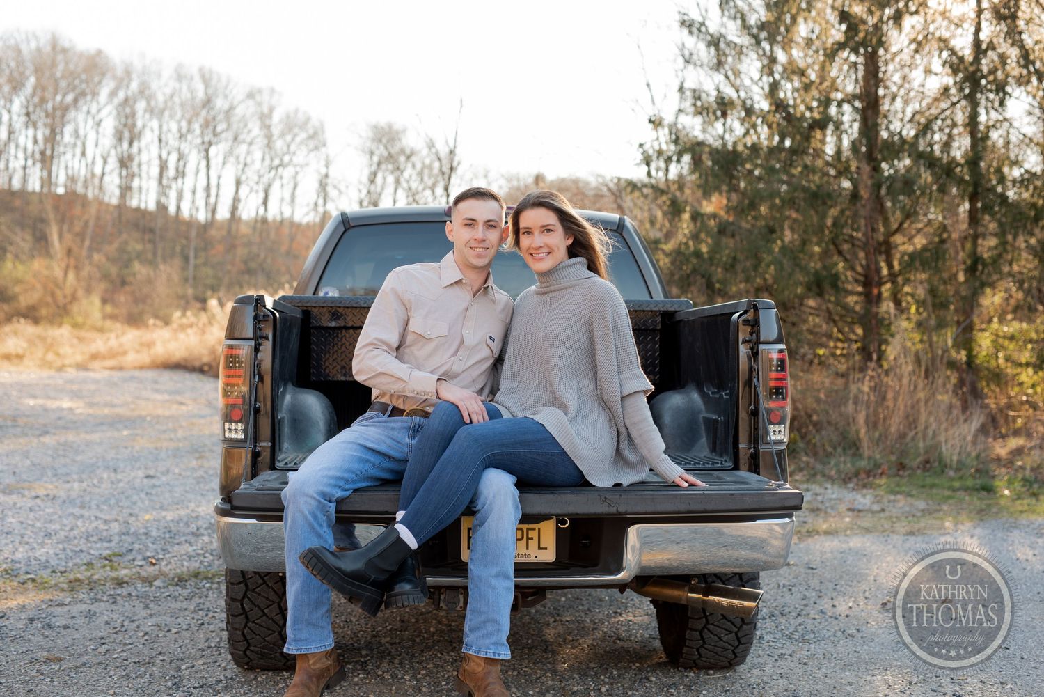 A couple sits together in casual attire on the tailgate of a black pickup truck at sunset.
