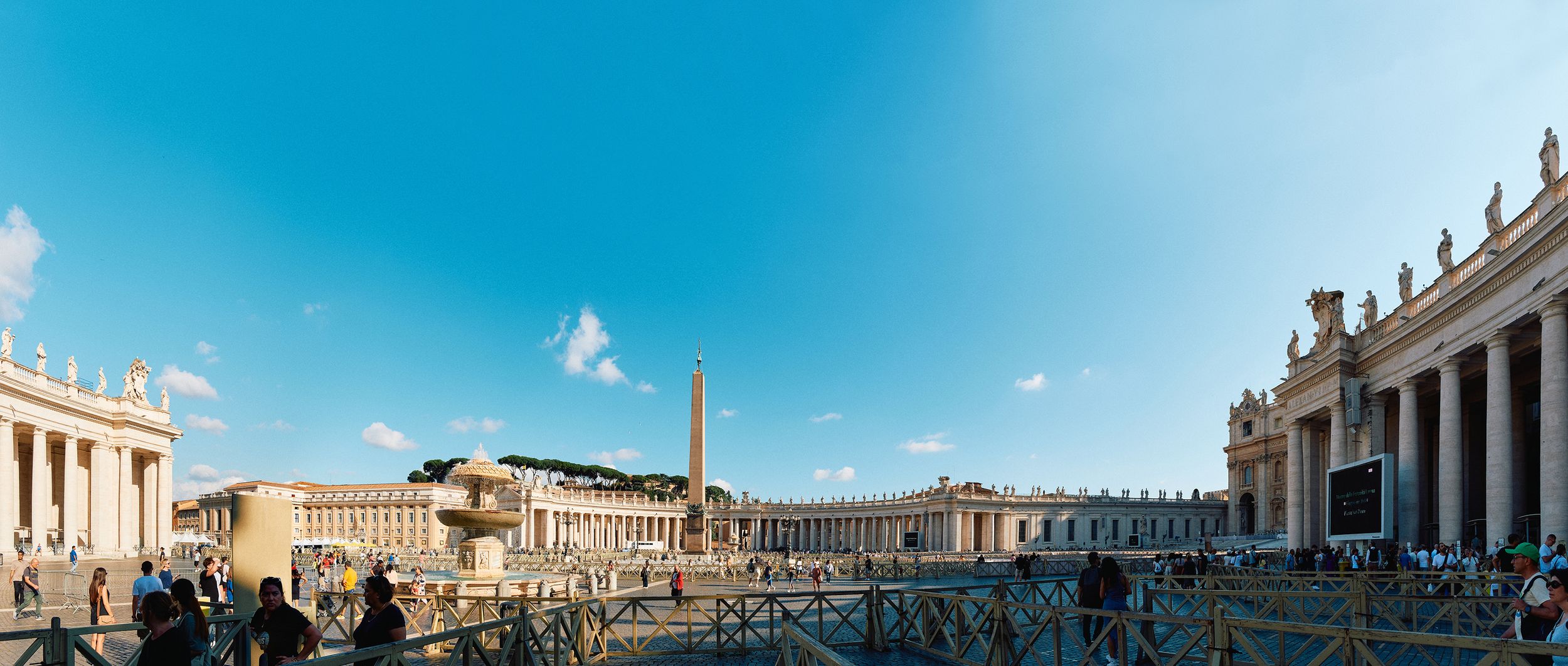 Panoramic view of Saint Peter's Square with obelisk and colonnades under bright blue sky.