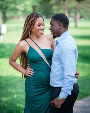 Romantic couple in emerald dress and blue shirt embrace while posing for engagement photos in a lush green park.