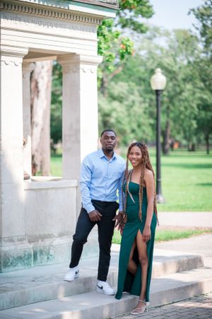 A couple poses together in a green dress and blue shirt near white columns outdoors.