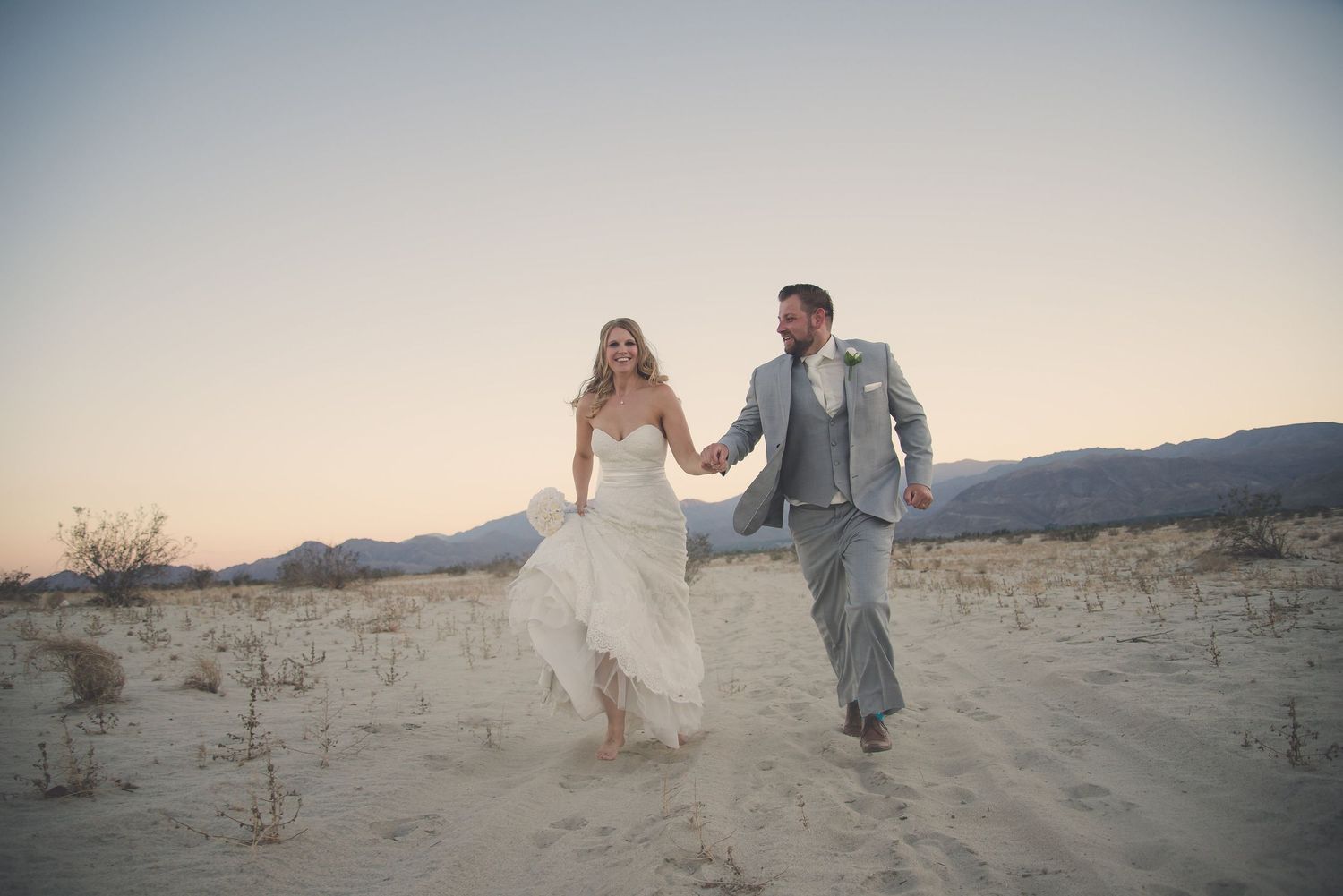 Bride and groom holding hands running through sandy desert landscape at sunset with mountains behind.