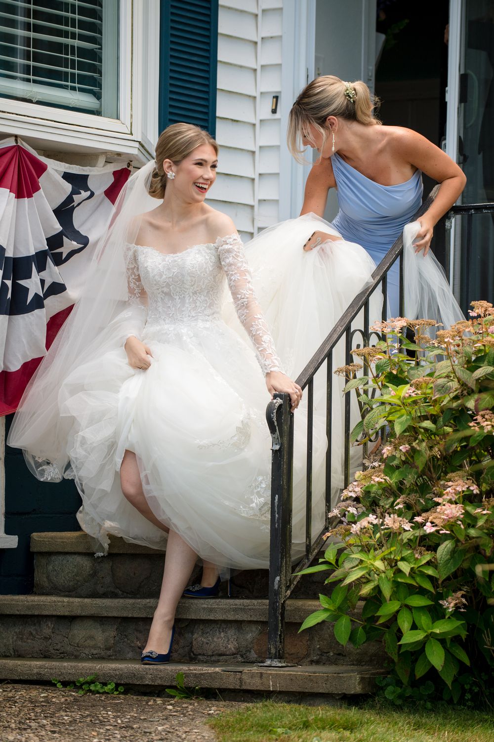 A bride in a lace wedding gown descends stairs with help from an attendant in a blue dress.