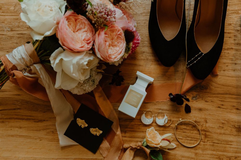 Wedding accessories including black heels, perfume bottle, floral bouquet and jewelry arranged on wooden floor. London Wedding.