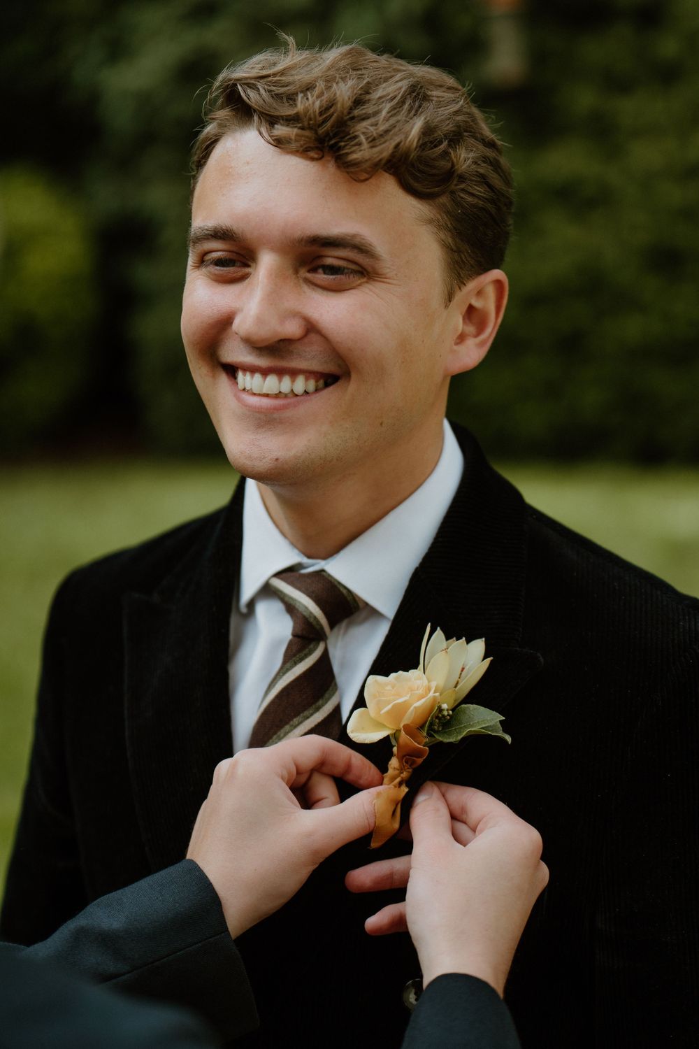 Someone pins a boutonniere onto a groom's lapel during an outdoor wedding. London Wedding.