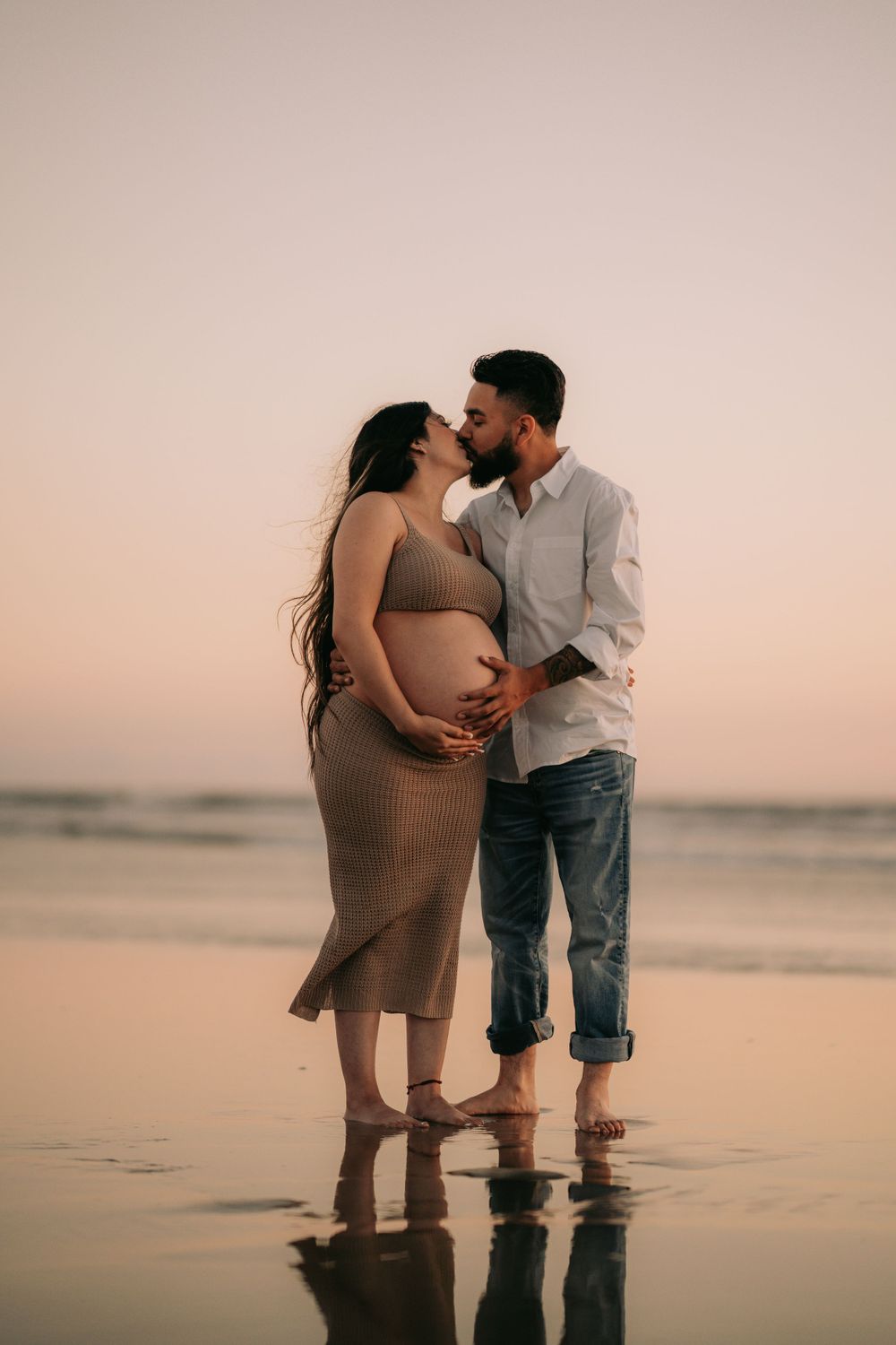 Expectant couple embracing on beach at sunset in romantic maternity photo.