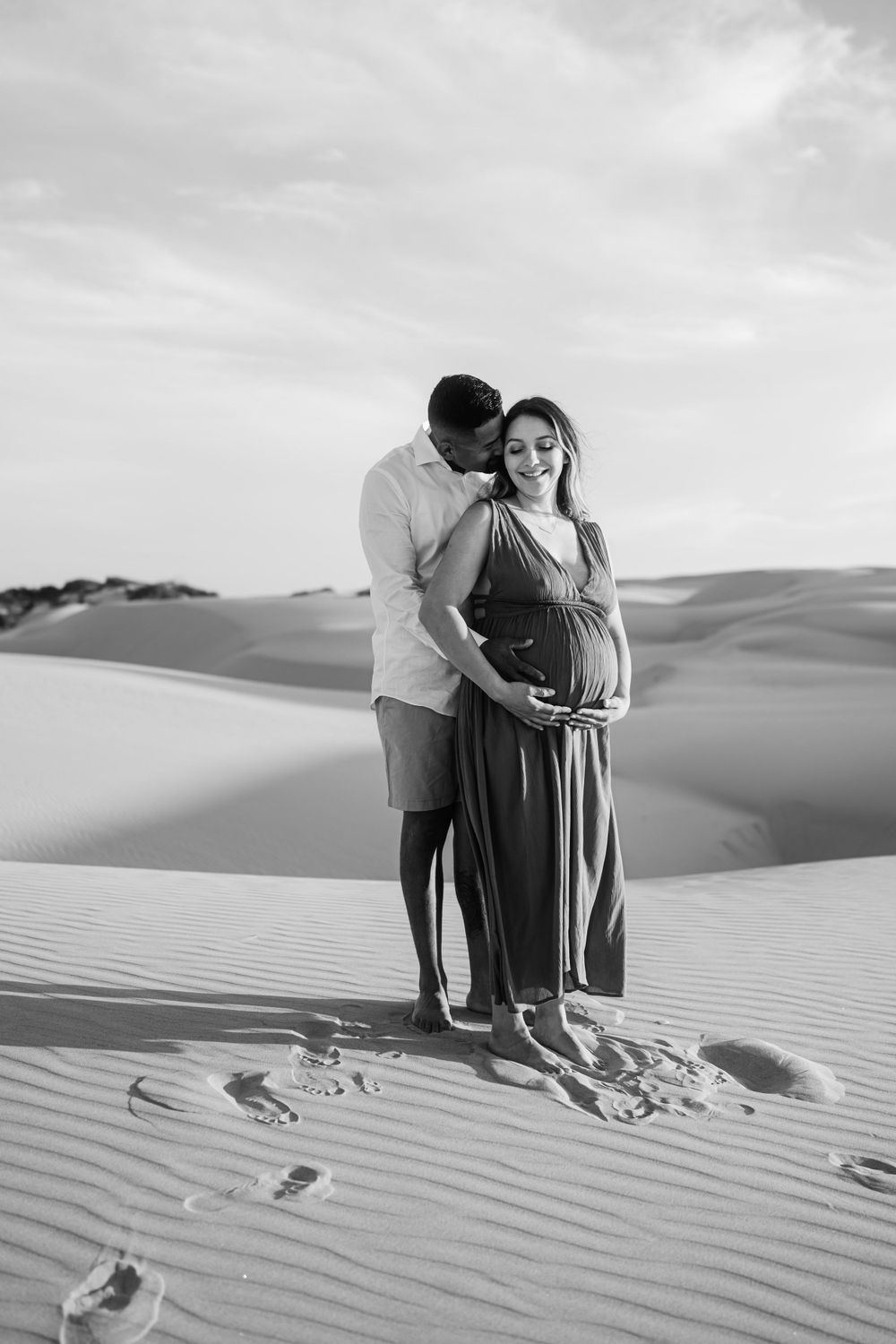 Couple shares intimate moment on sand dunes in dramatic black and white photography.
