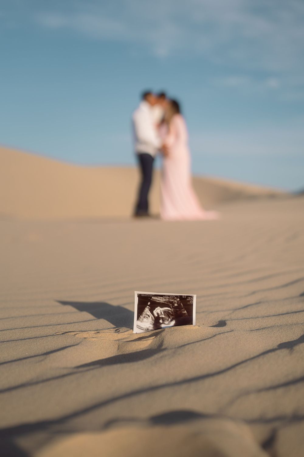 Ultrasound photo in sand with blurred couple silhouette against desert dunes backdrop.