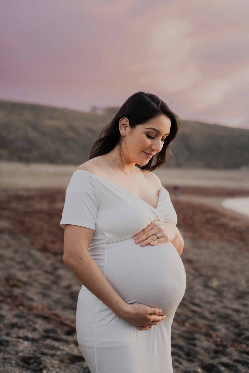 Expectant mother in white dress cradling baby bump during sunset beach photoshoot.