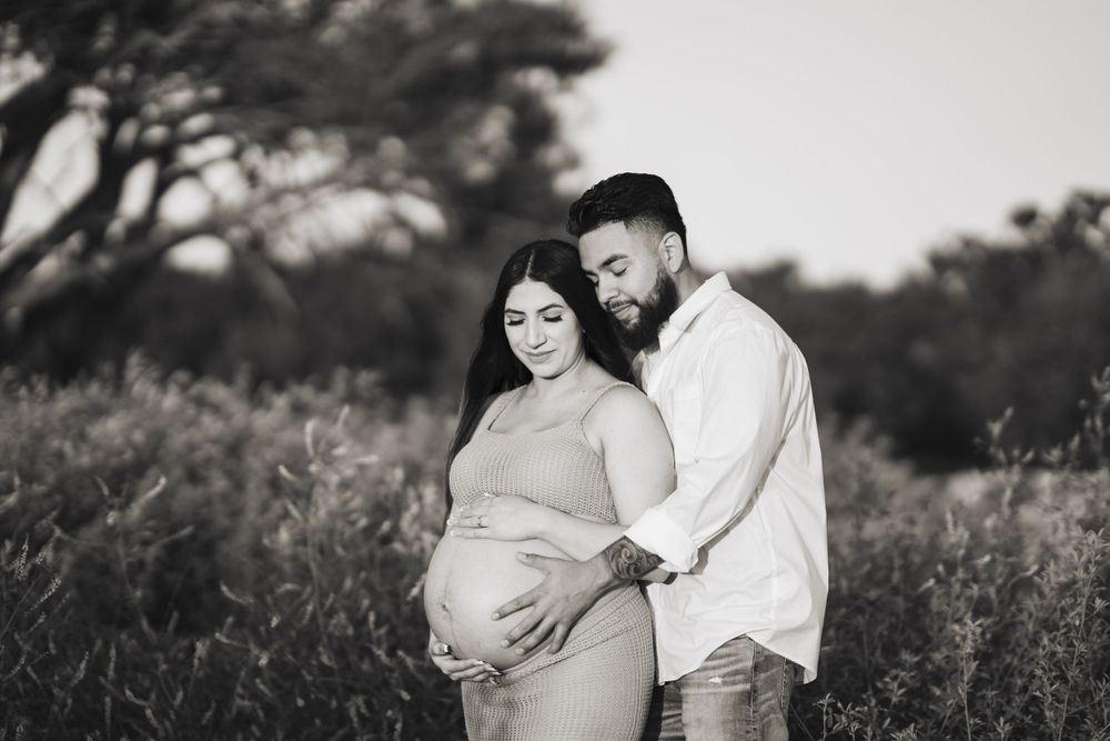Black and white maternity photo of couple embracing in field with trees.