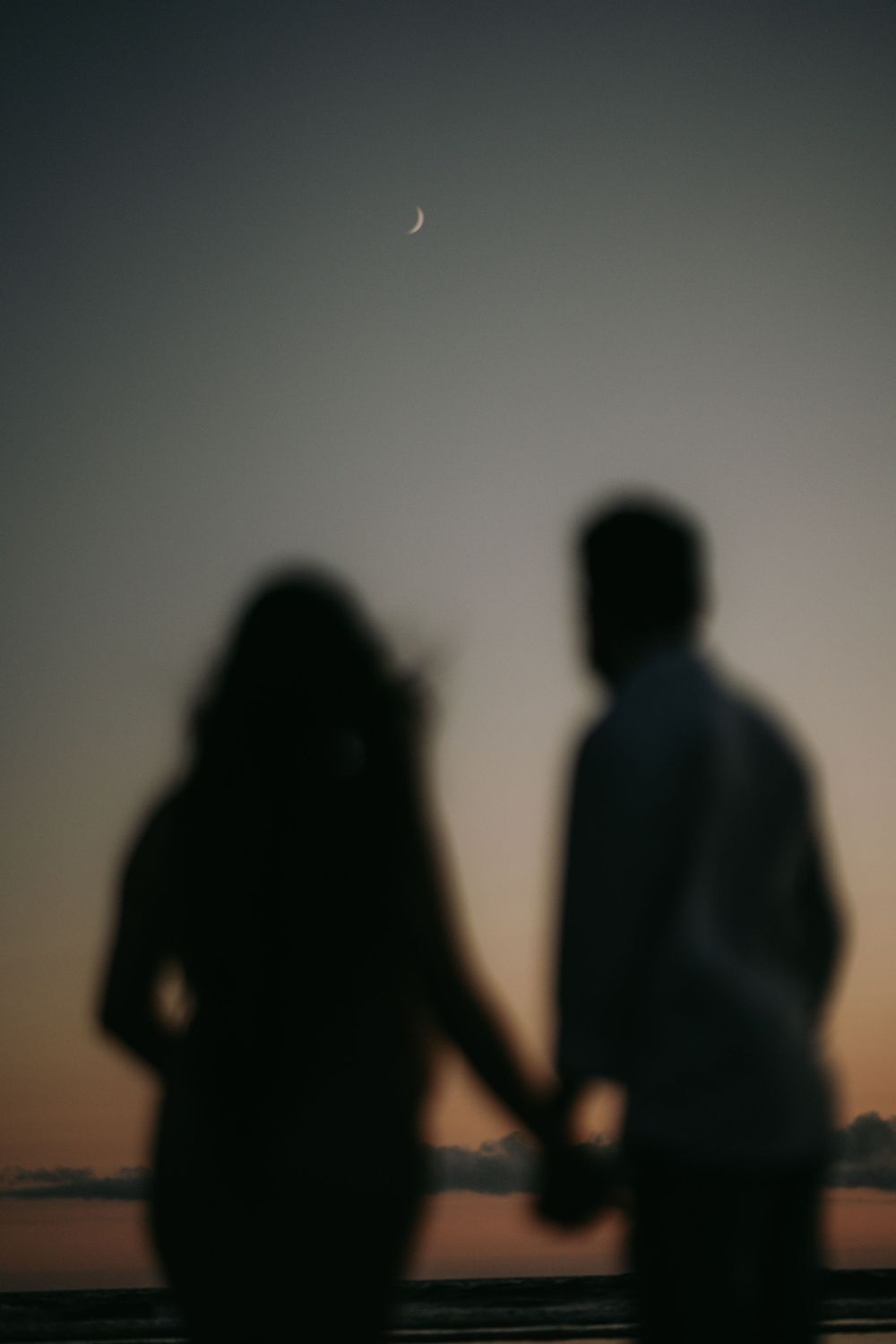 Silhouetted couple holding hands against twilight sky with crescent moon.