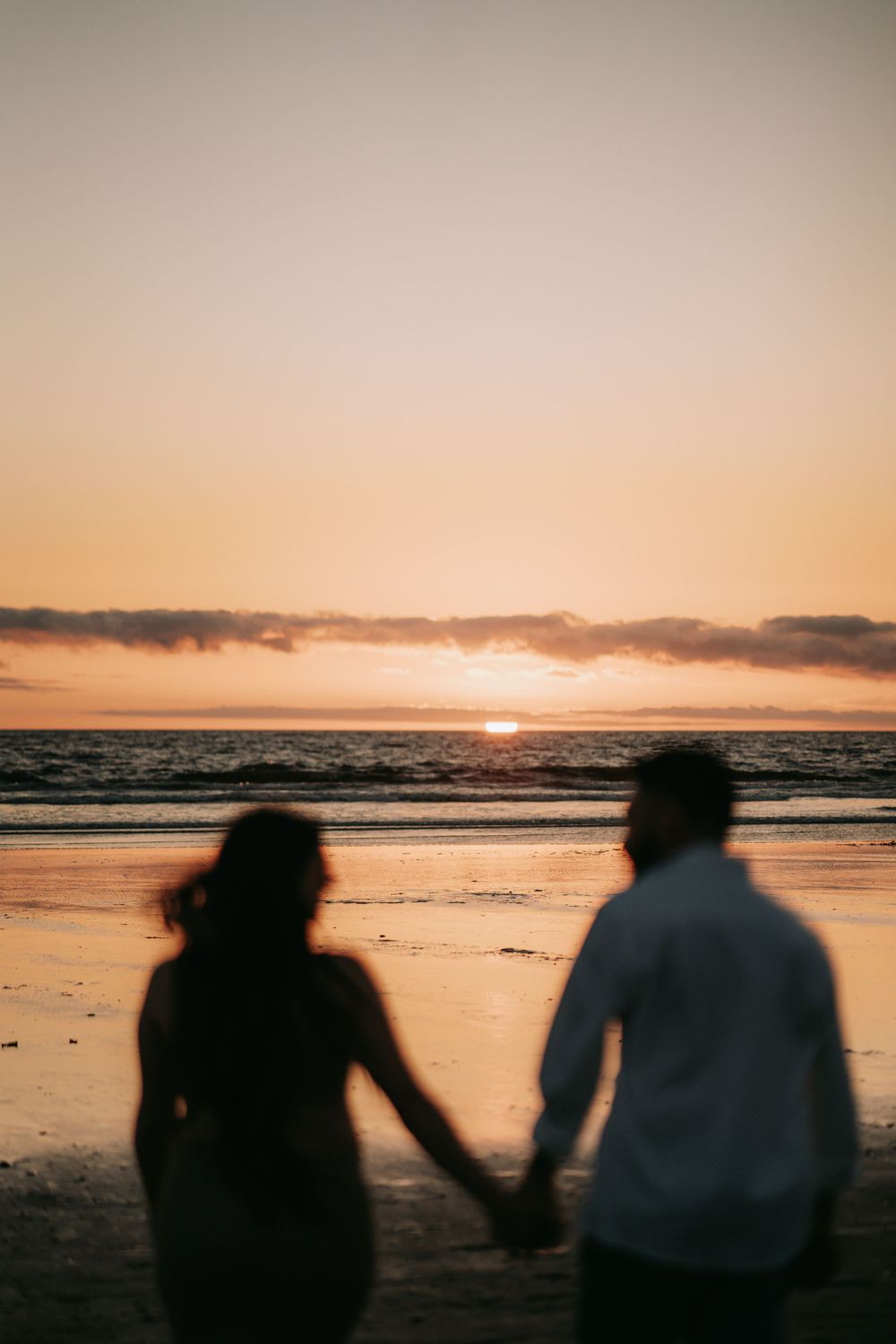 Couple silhouetted against orange sunset sky holding hands on beach.