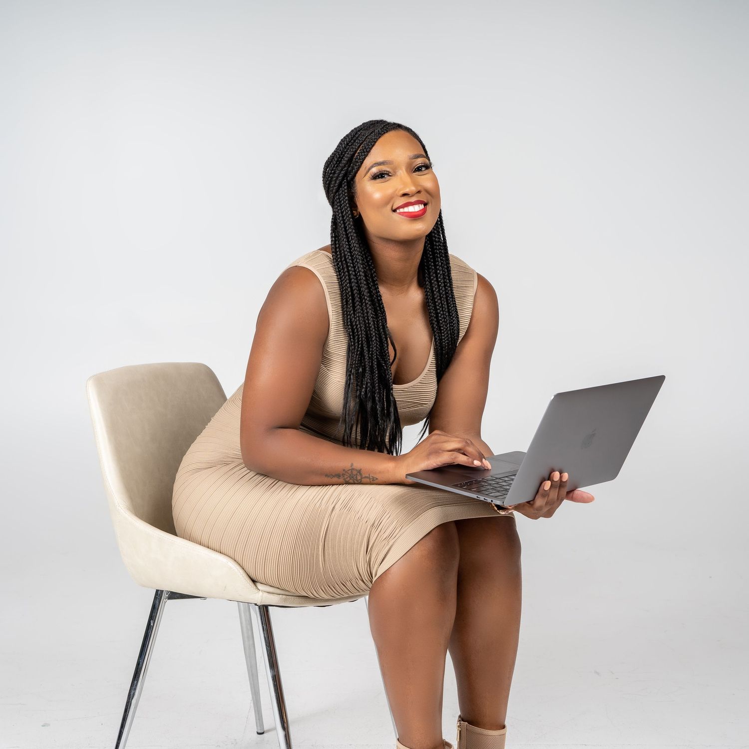 Woman in beige dress sits with laptop, smiling against white studio background.