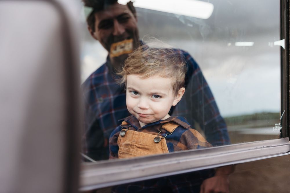 A young child in brown overalls smiles through a car window while someone watches from behind.