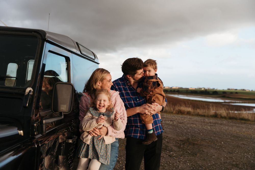 A family stands together by their black vehicle on an overcast day in a rural setting.