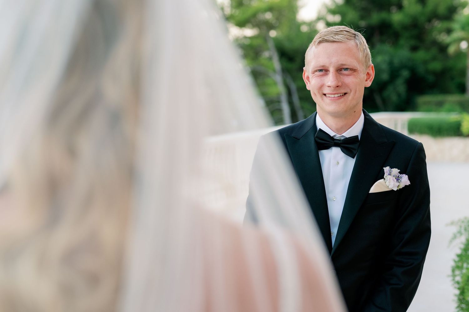 Groom in classic black tuxedo smiles during outdoor wedding ceremony.