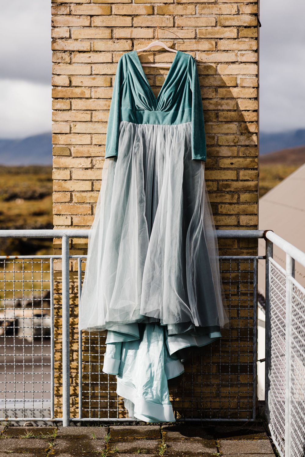 Light blue and white wedding dress hanging on brick wall against mountain landscape.