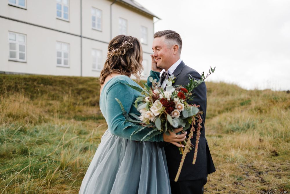 Couple shares an intimate moment as bride holds autumn-inspired bouquet on grassy hillside.