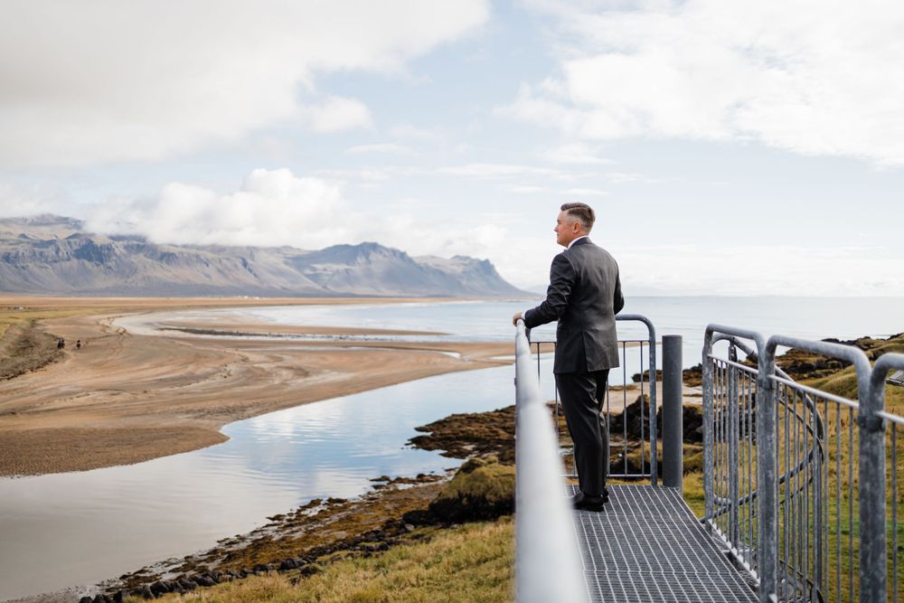 Person standing at metal railing overlooking scenic coastal landscape with mountains and water.