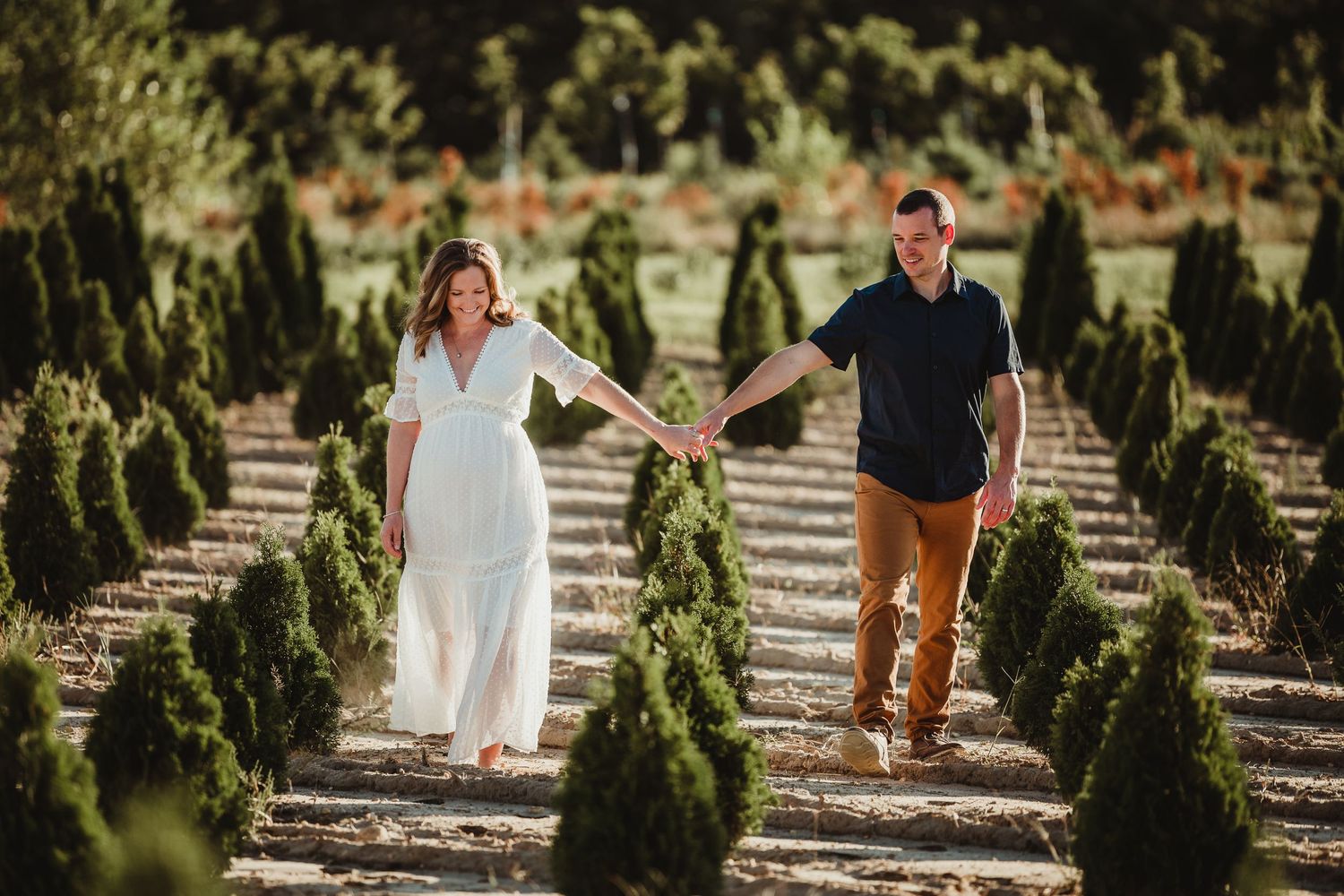 Couple walking hand in hand through rows of small evergreen trees in a Christmas tree farm.