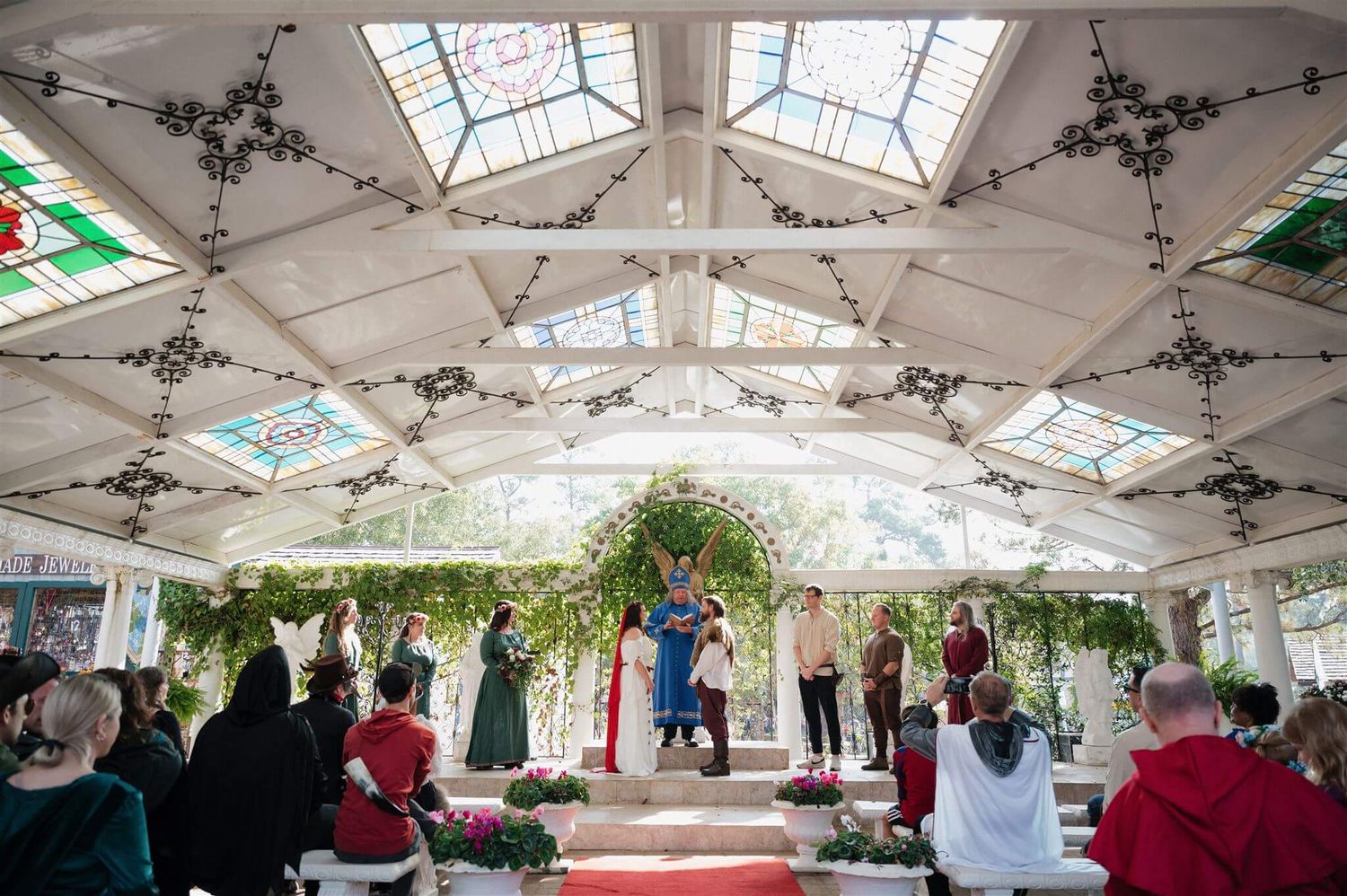 Wedding ceremony under ornate pavilion with stained glass ceiling and decorative ironwork.