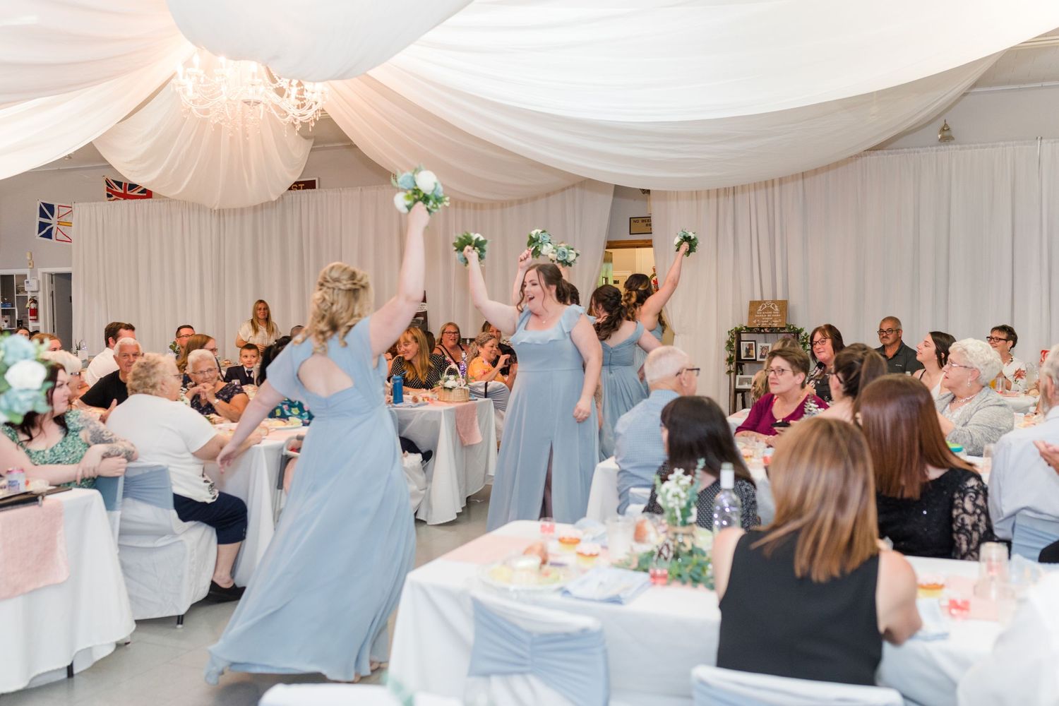 Wedding reception guests in light blue bridesmaid dresses dancing and celebrating in a white draped venue.