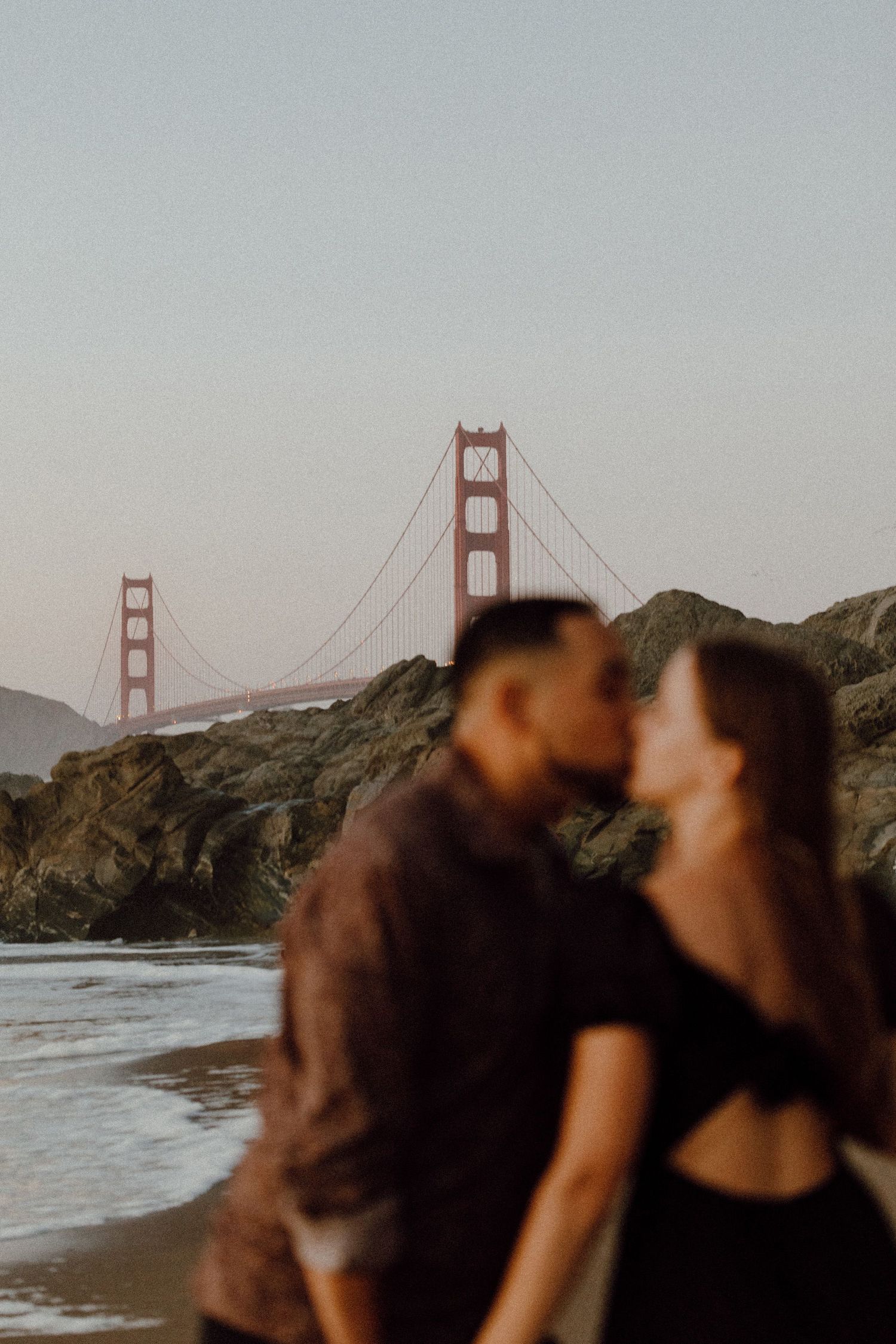 A couple kisses with San Francisco's Golden Gate Bridge in the background behind them. Couple is out of focus & grainyai