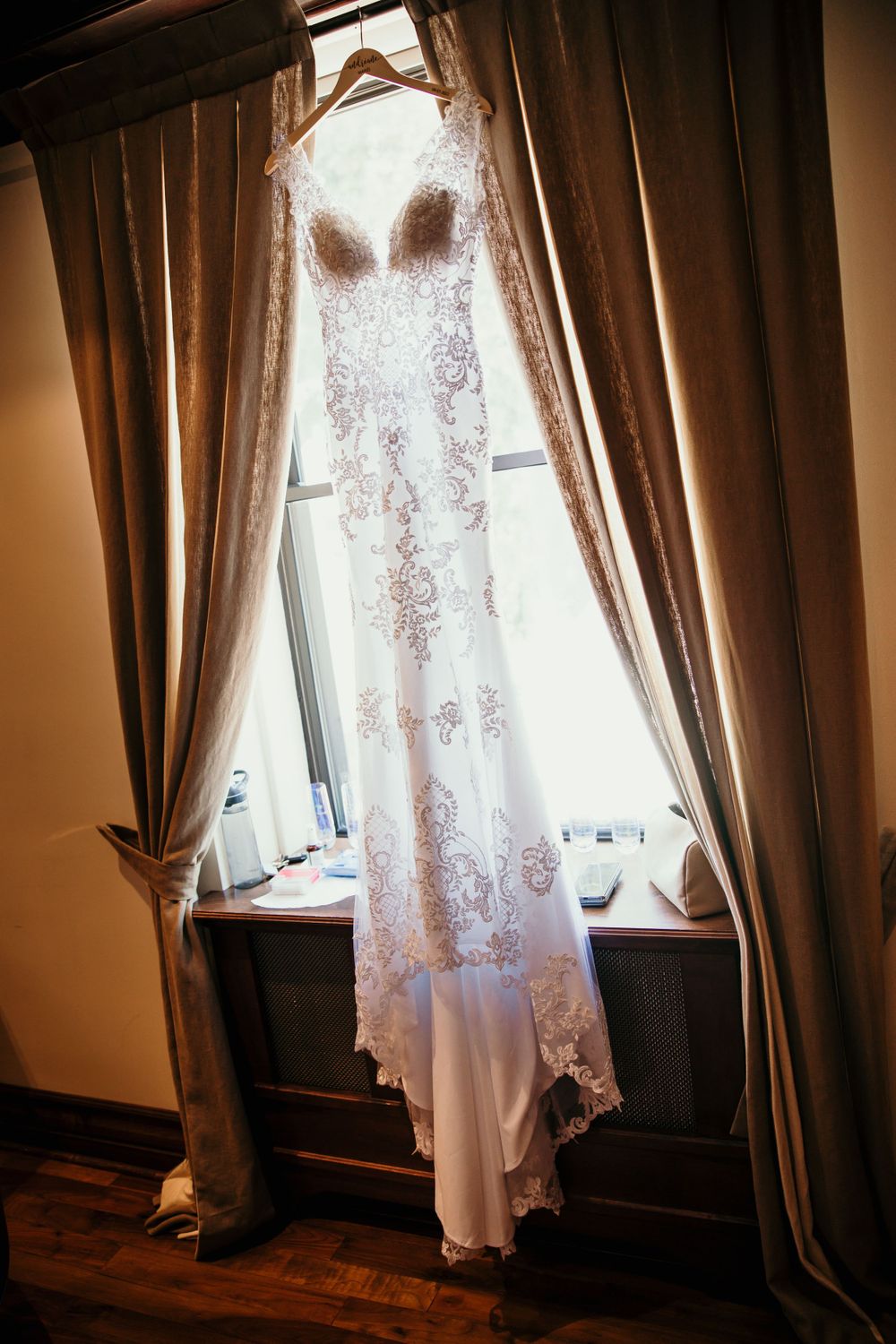 An elegant lace wedding dress hangs in a window frame with dramatic curtains.