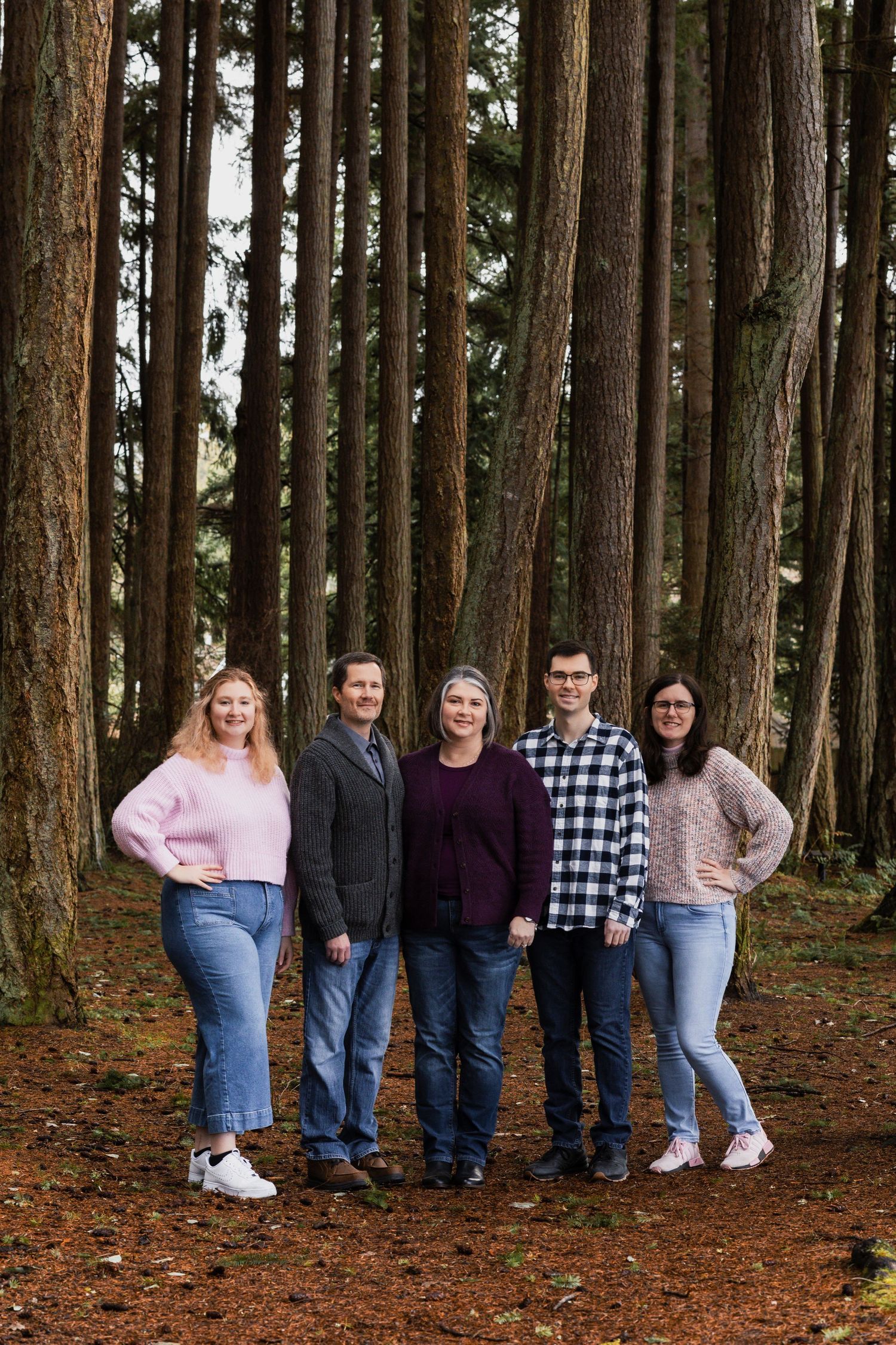A group portrait of five people standing among tall evergreen trees in a forest.