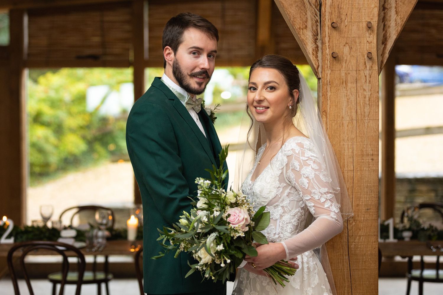 A bride and groom embrace each other while looking side on to the camera. Posed against a wooden beam inside Cripps Barn