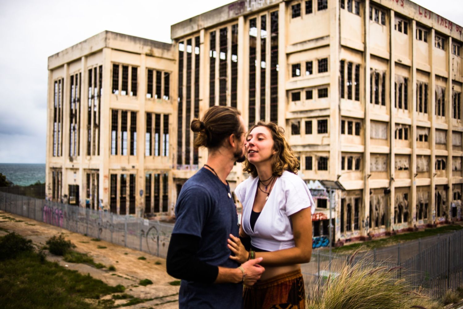 A couple embraces in front of an abandoned industrial building.
