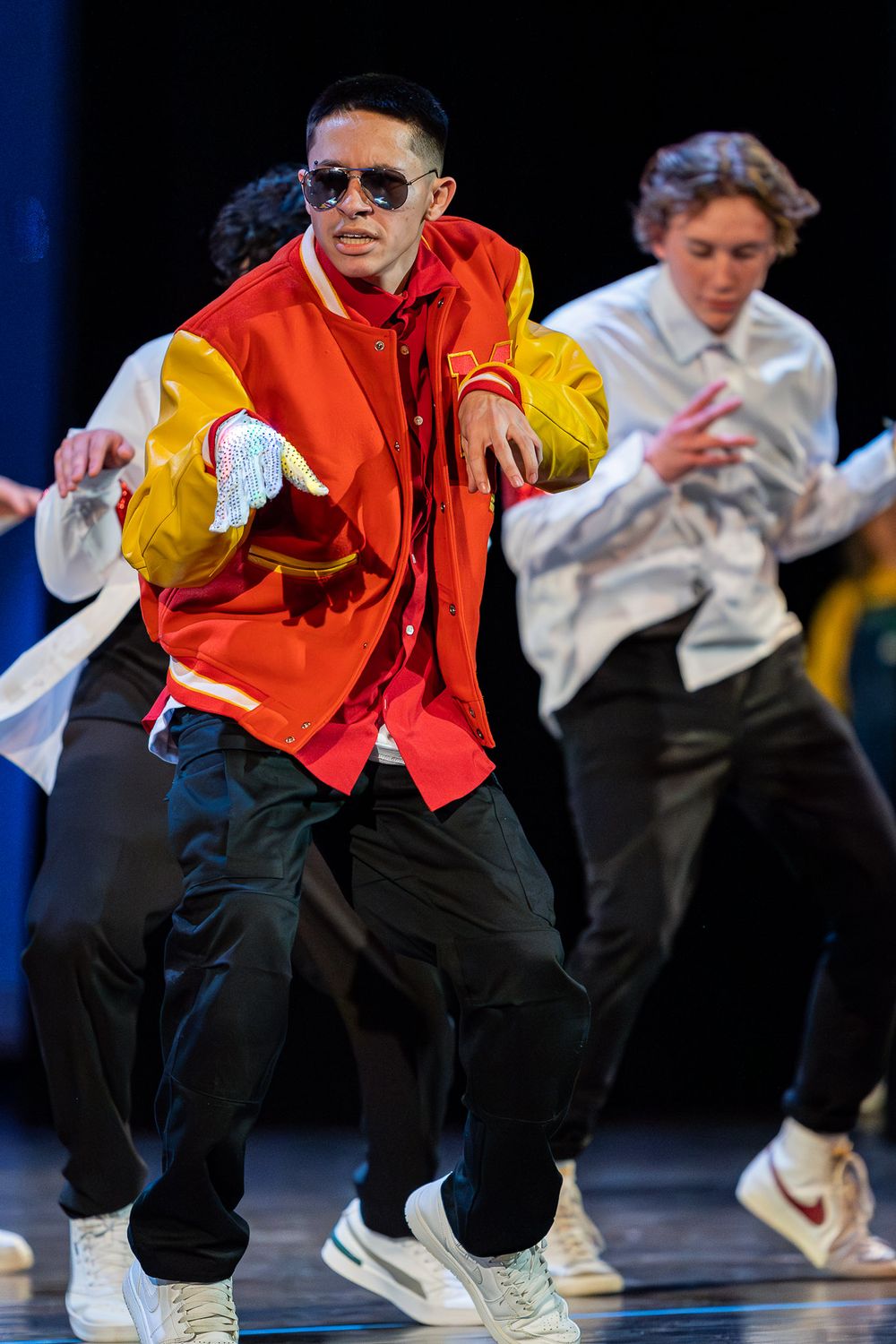 Dancer in bright red and yellow jacket performs hip hop routine on dark stage.