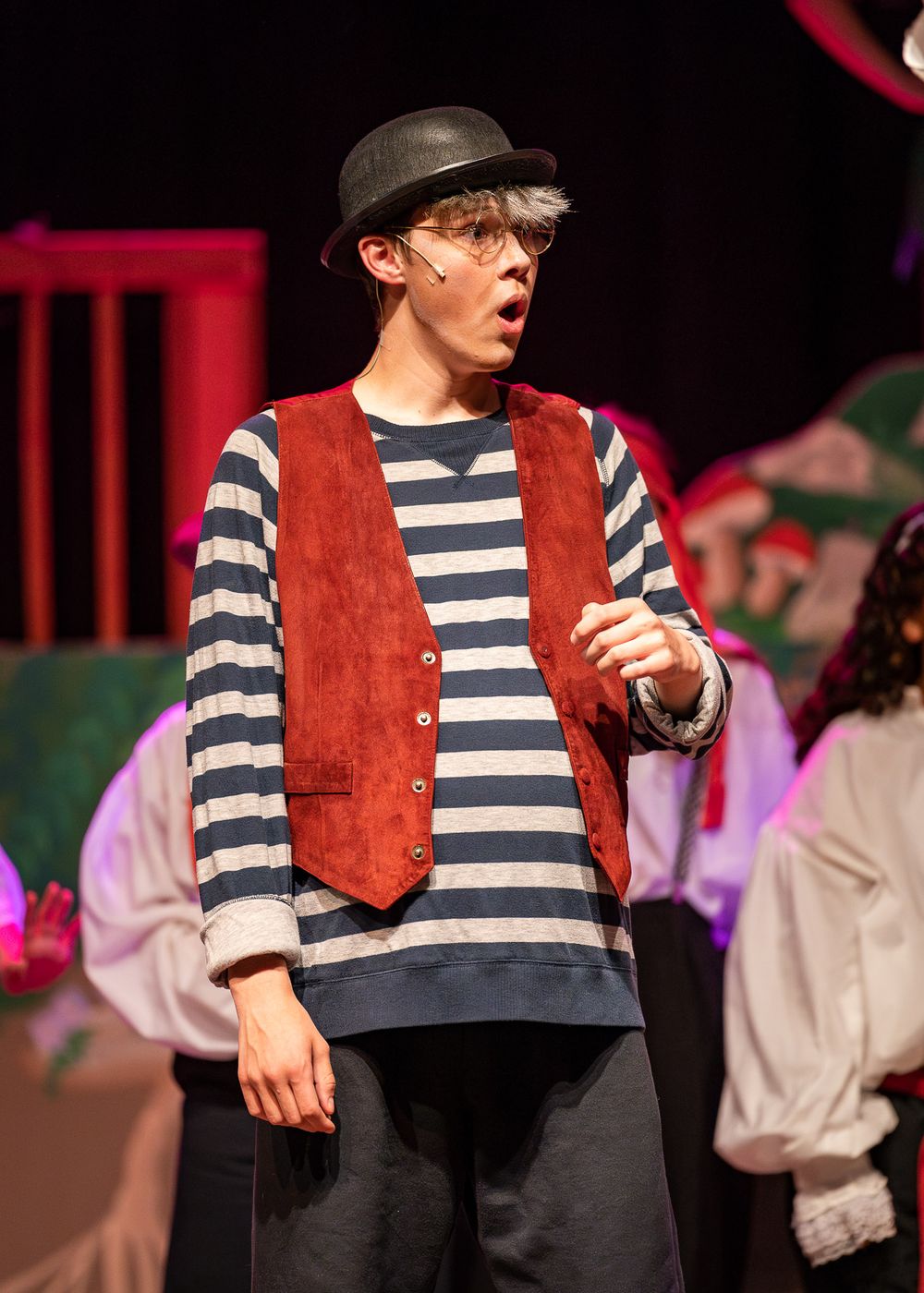 A performer wearing a striped shirt, vest and bowler hat acts expressively on stage.