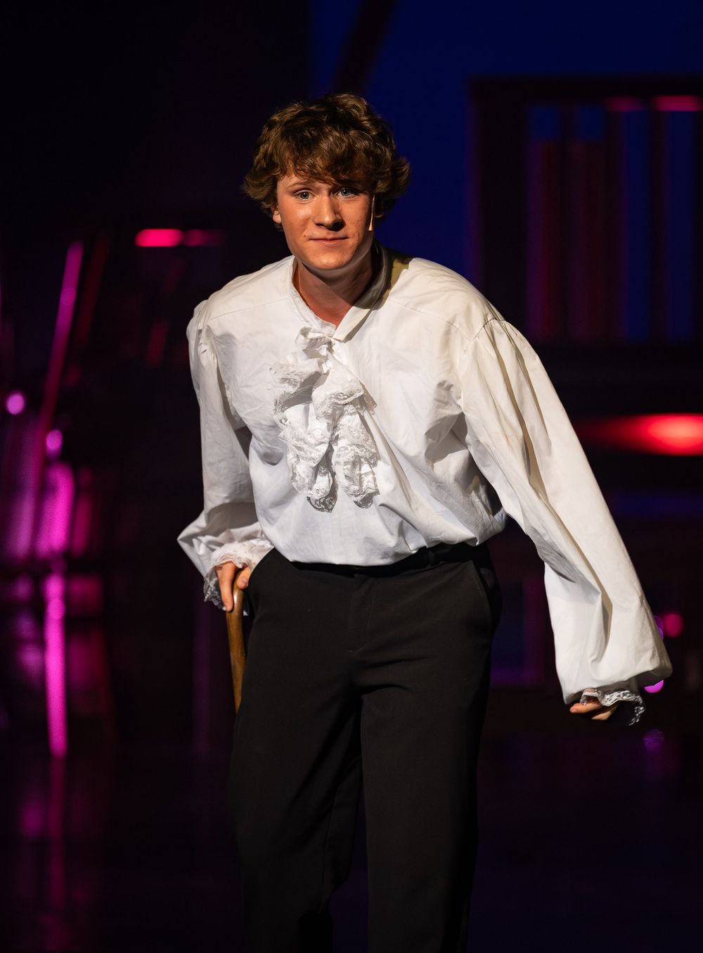 A performer in a white ruffled shirt poses dramatically on a dimly lit stage.