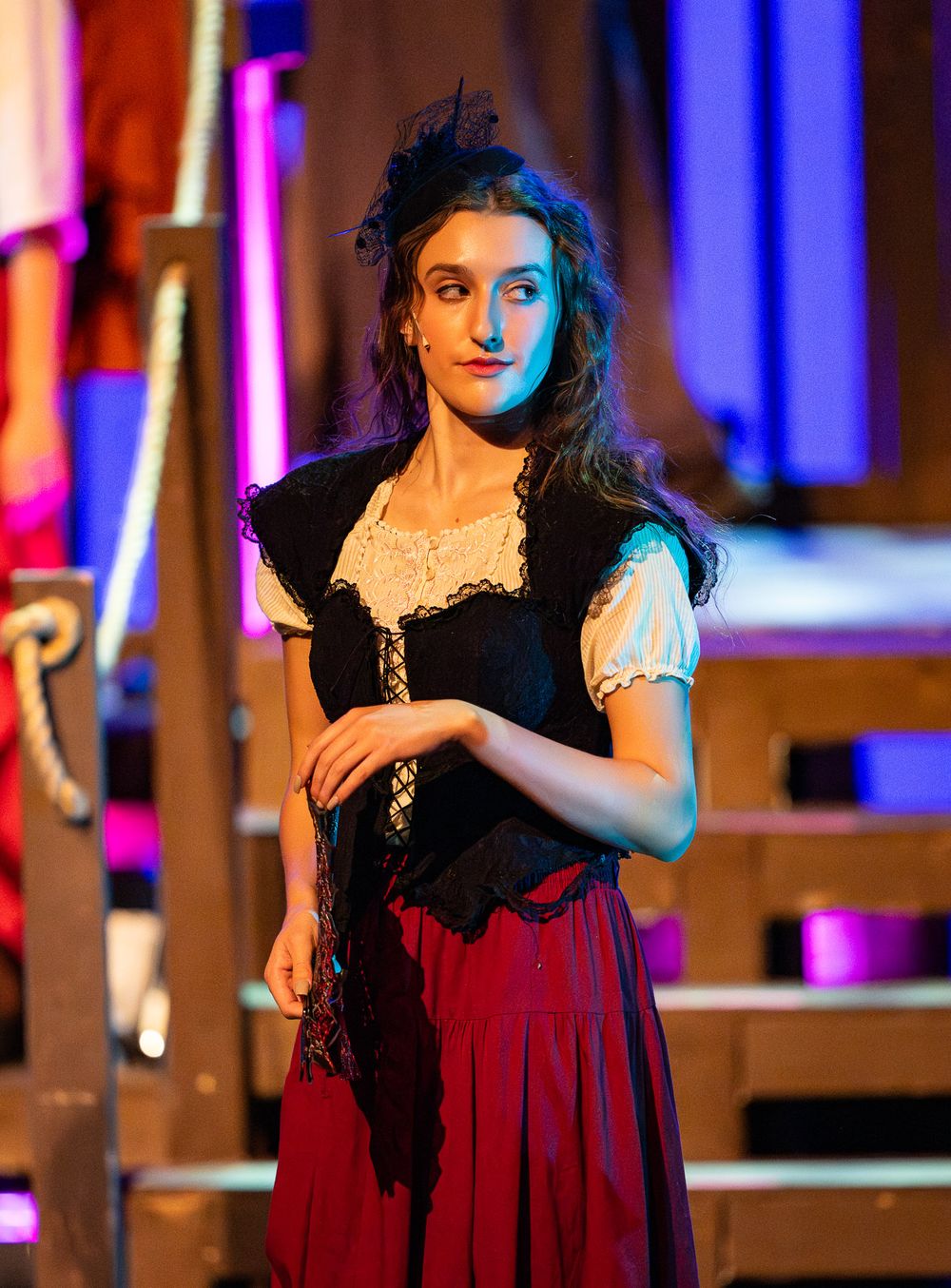 Actress in period costume with black corset and red skirt performing on stage with dramatic lighting.