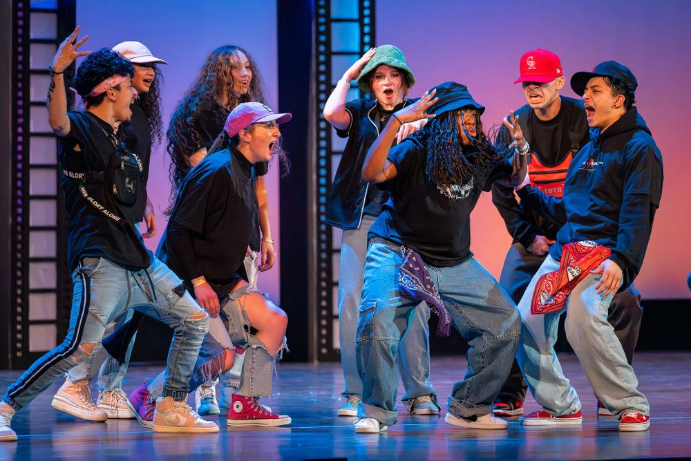 Group of street dancers performing choreographed hip hop routine on stage with colored lighting.