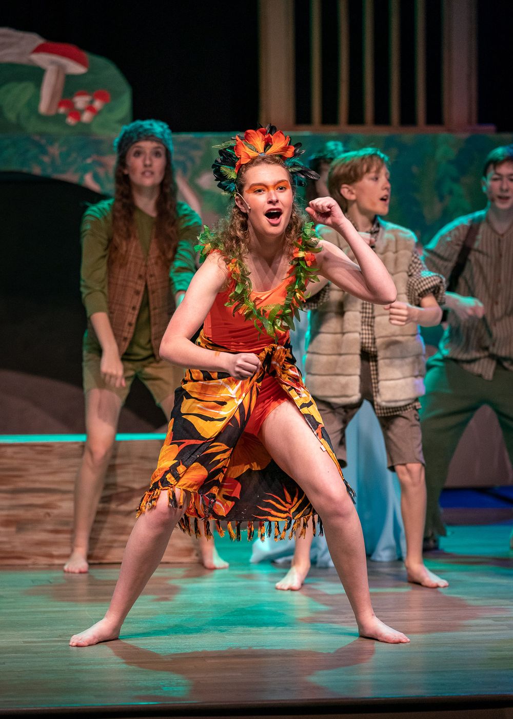 Performer in tribal costume dancing energetically on stage with tropical scenery backdrop.